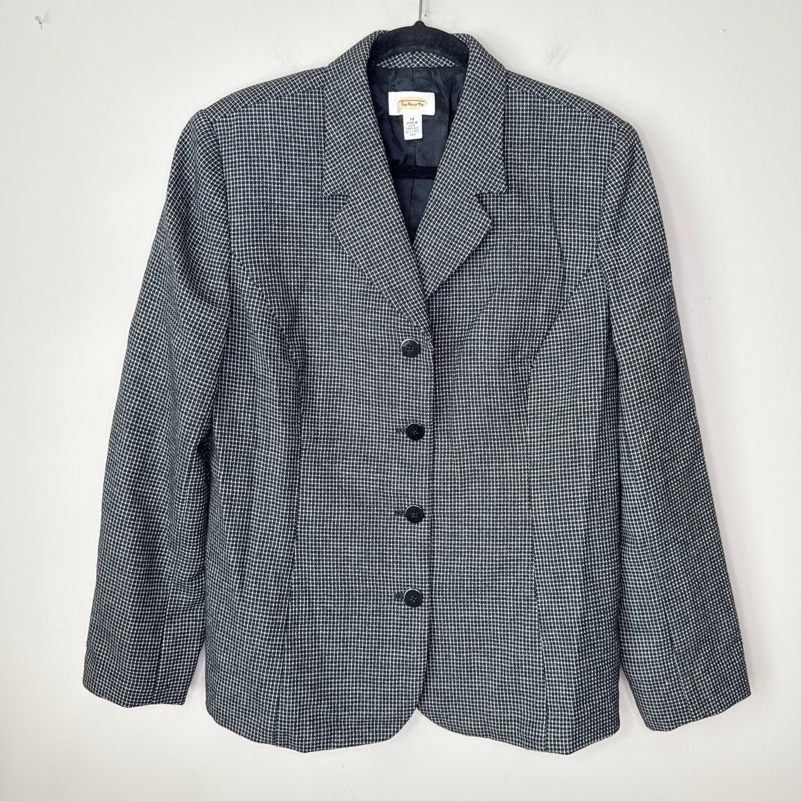 Black and white houndstooth blazer with notch lapel hanging against a white wall.