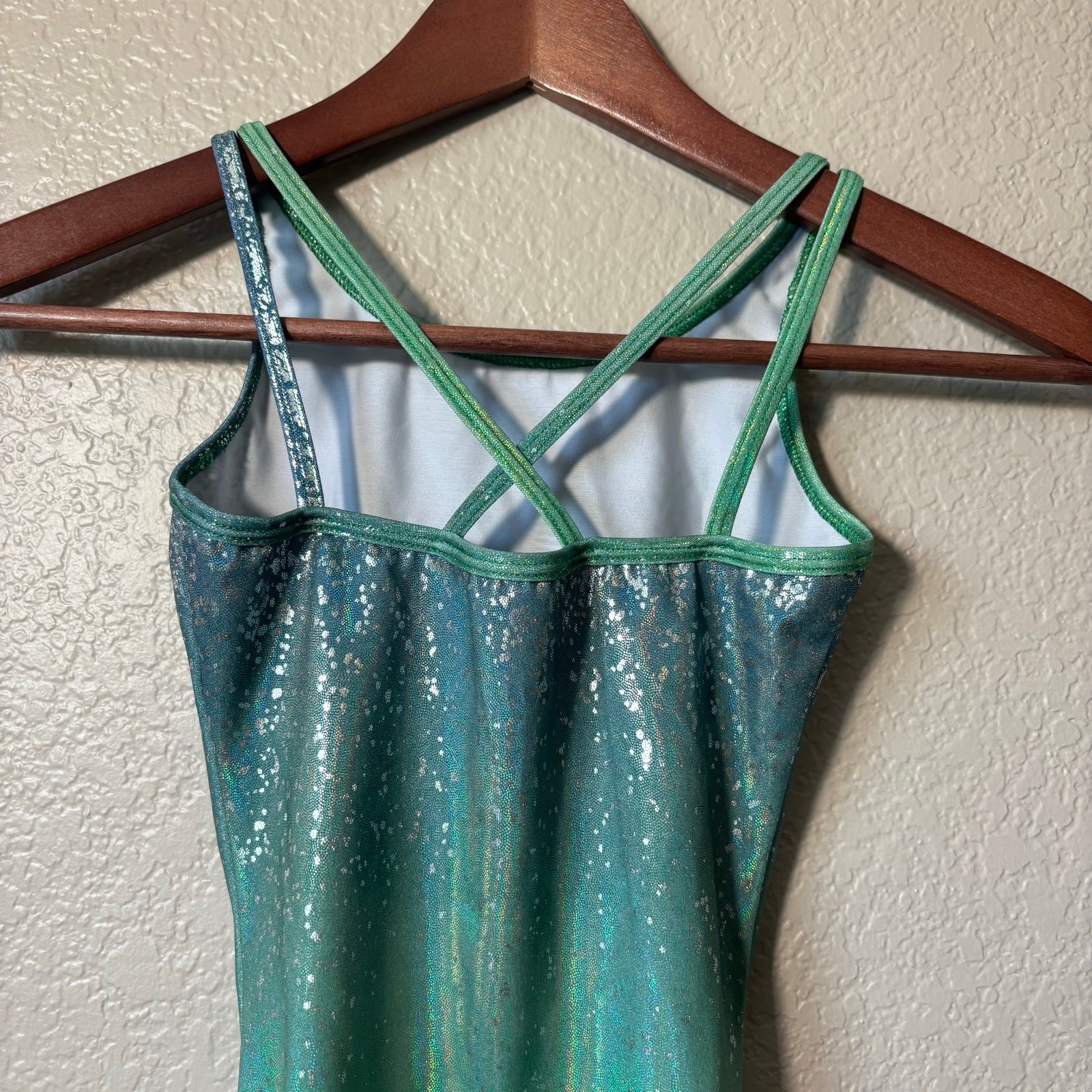 Green and silver holographic Destira gymnastics leotard with thin straps and scalloped lace trim on wooden hanger.