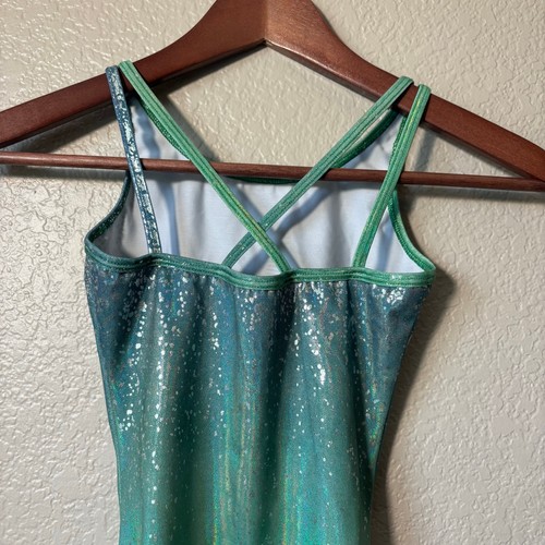 Green and silver holographic Destira gymnastics leotard with thin straps and scalloped lace trim on wooden hanger.