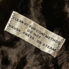White fabric tag on brown fur coat with red “CLEAN BY FUR COAT METHOD” text, attached by silver zigzag stitching.