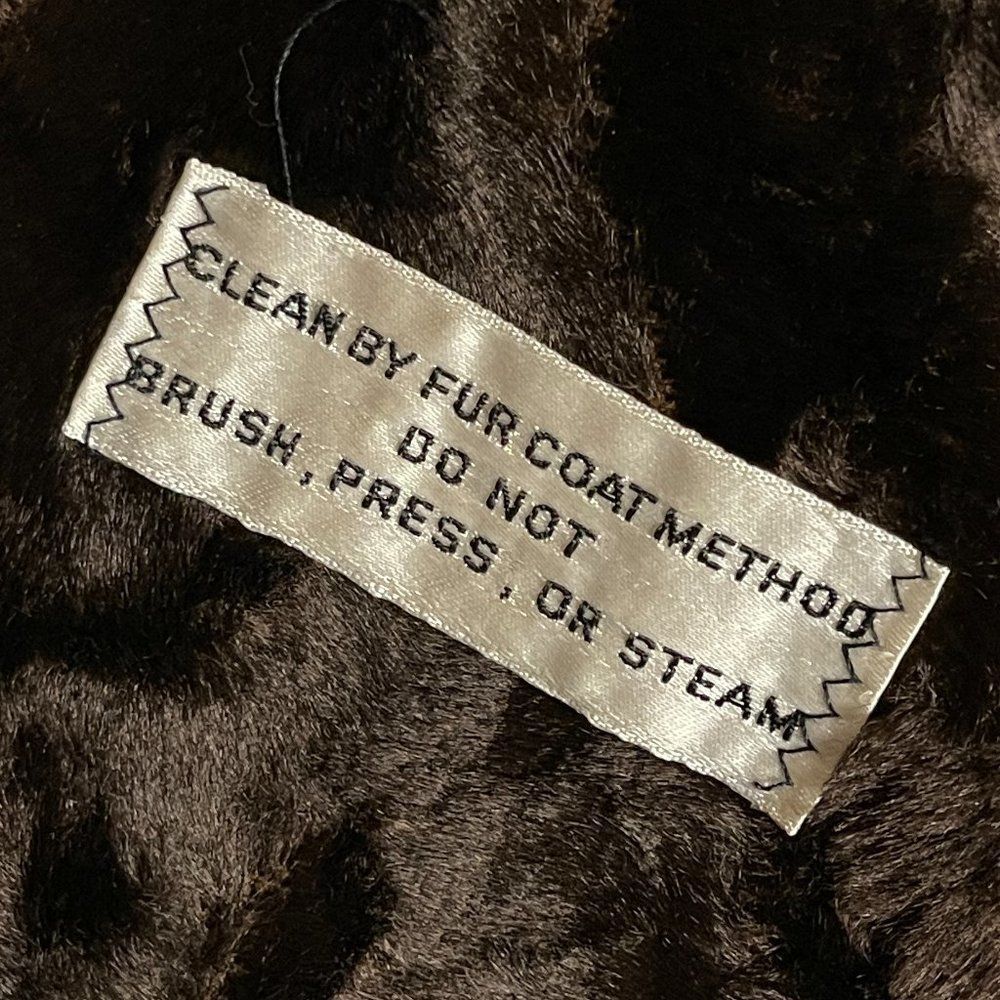 White fabric tag on brown fur coat with red “CLEAN BY FUR COAT METHOD” text, attached by silver zigzag stitching.