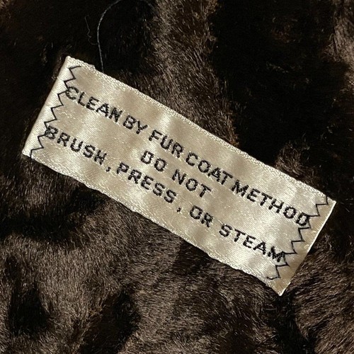 White fabric tag on brown fur coat with red “CLEAN BY FUR COAT METHOD” text, attached by silver zigzag stitching.