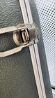 Weathered silver metal strap with worn brass hinge against green textured hard case on light grid background.