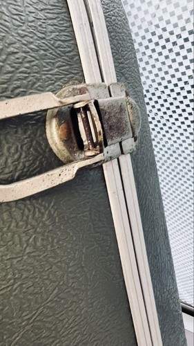 Weathered silver metal strap with worn brass hinge against green textured hard case on light grid background.