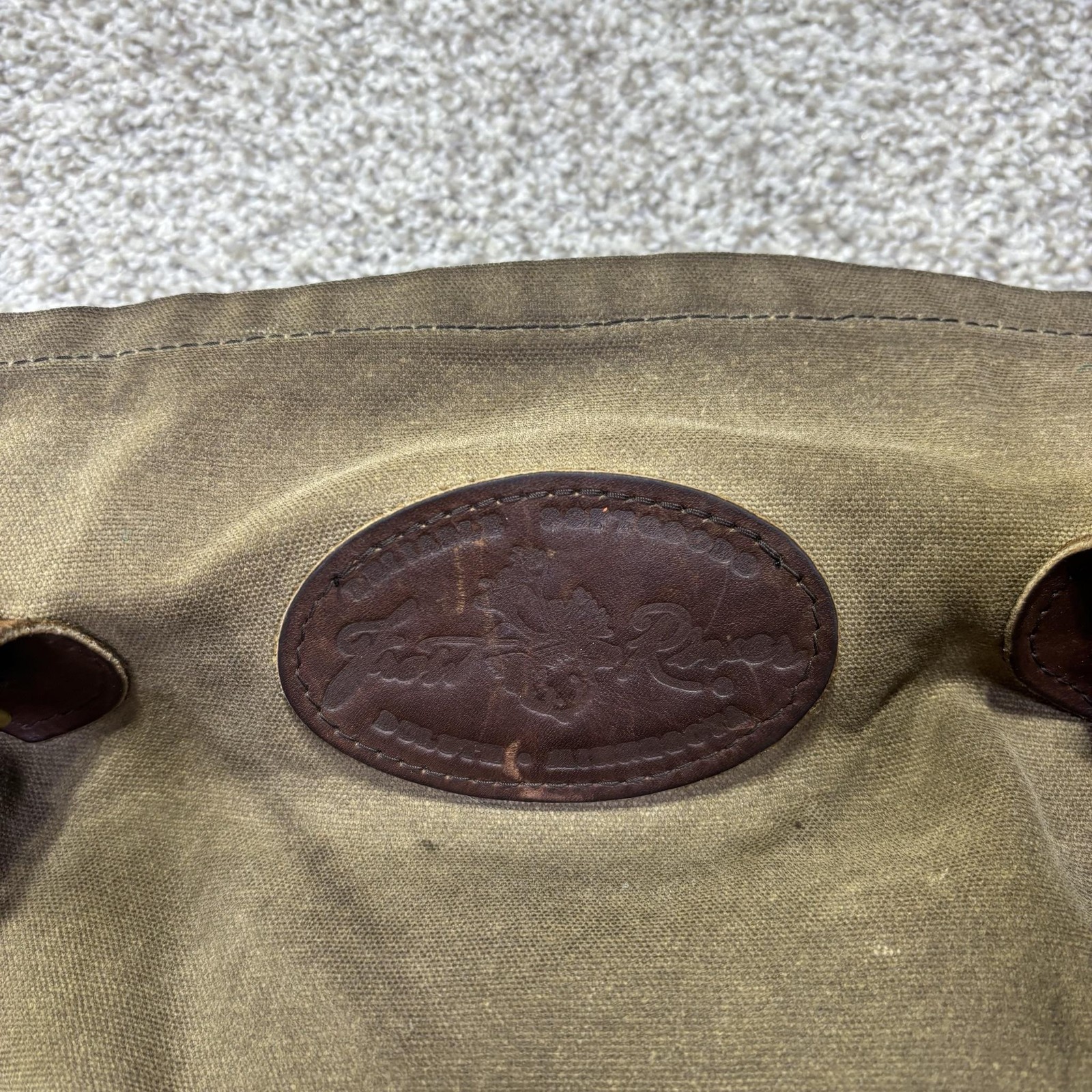 Brown canvas crossbody bag with dark brown leather strap, featuring embossed logo patch on front.