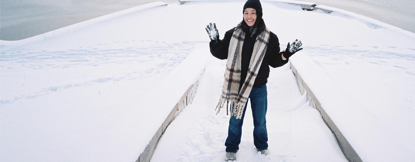 Eine Person trägt Boyfriend Jeans, eine Winterjacke, einen Schal, Mütze und Handschuhe und steht lächelnd im Schnee auf einem verschneiten Weg.