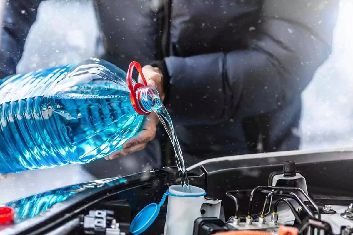 A driver wearing a winter coat pours blue fluid into a car’s windshield washer reservoir, as the snow falls.