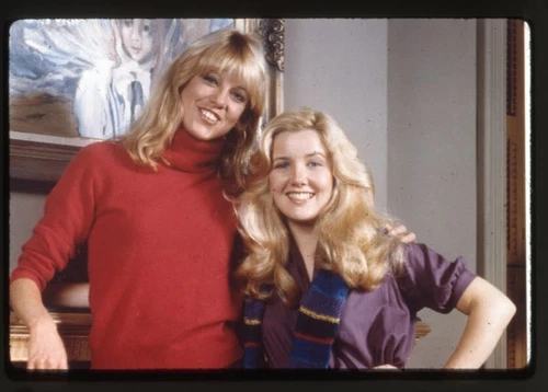 Roberta Leighton and Melody Thomas in red and purple clothing with blonde hair, standing in front of a framed painting.