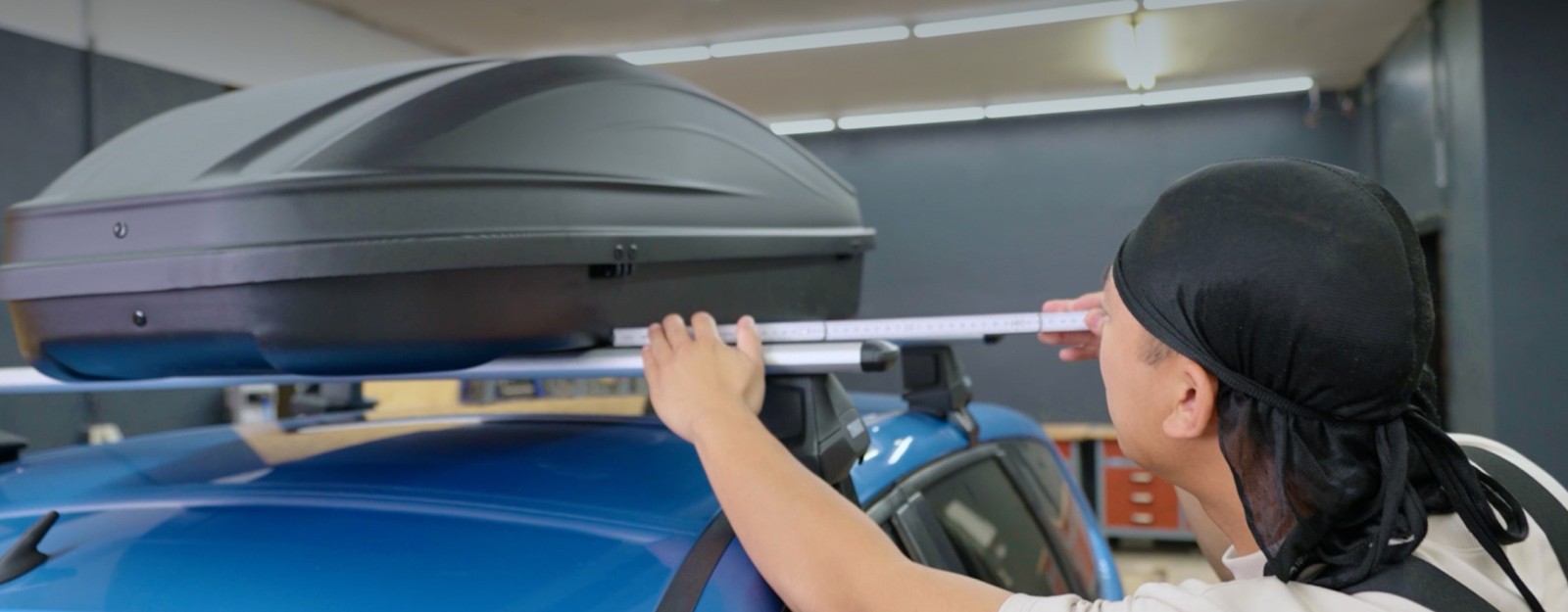 Aufnahme eines Mannes, der auf dem Dach eines blauen Autos eine schwarze Dachbox mit einem Zollstock ausmisst.