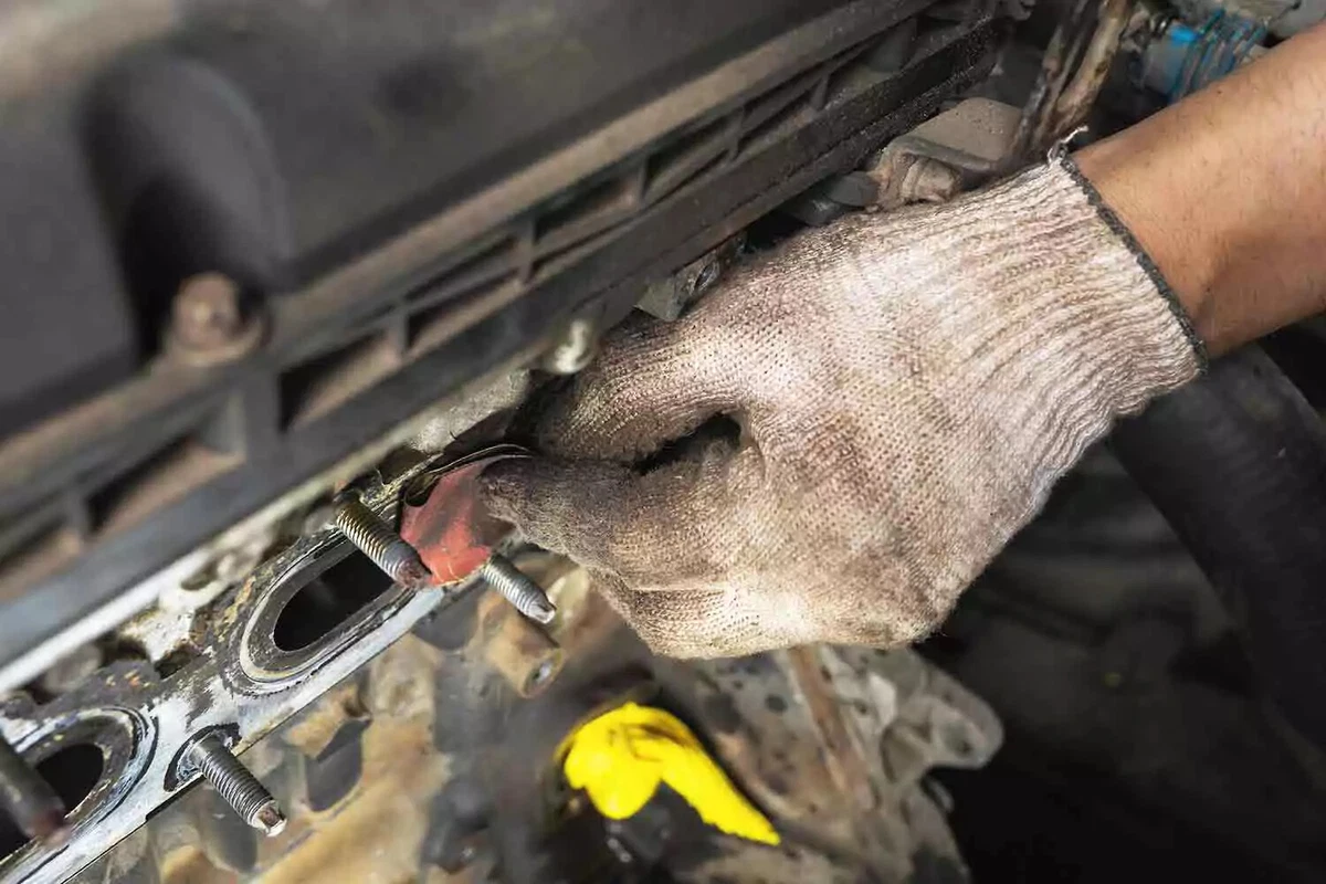 A mechanic wearing a white knit glove removes the old gasket residue on a cylinder head before installing a new exhaust manifold gasket.