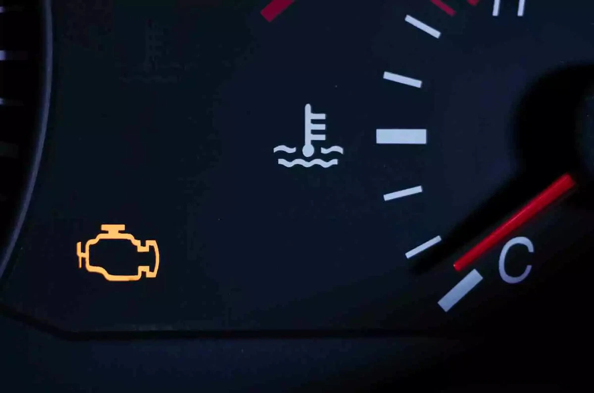 Close-up view of an illuminated check engine light on a vehicle’s dashboard, alongside the engine temperature gauge.