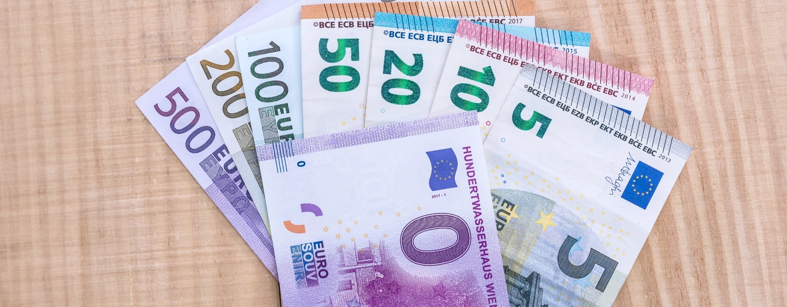 Aufnahme von einem 0-Euro-Schein, der über allen anderen verfügbaren Euro-Scheinen auf einem Holztisch liegt