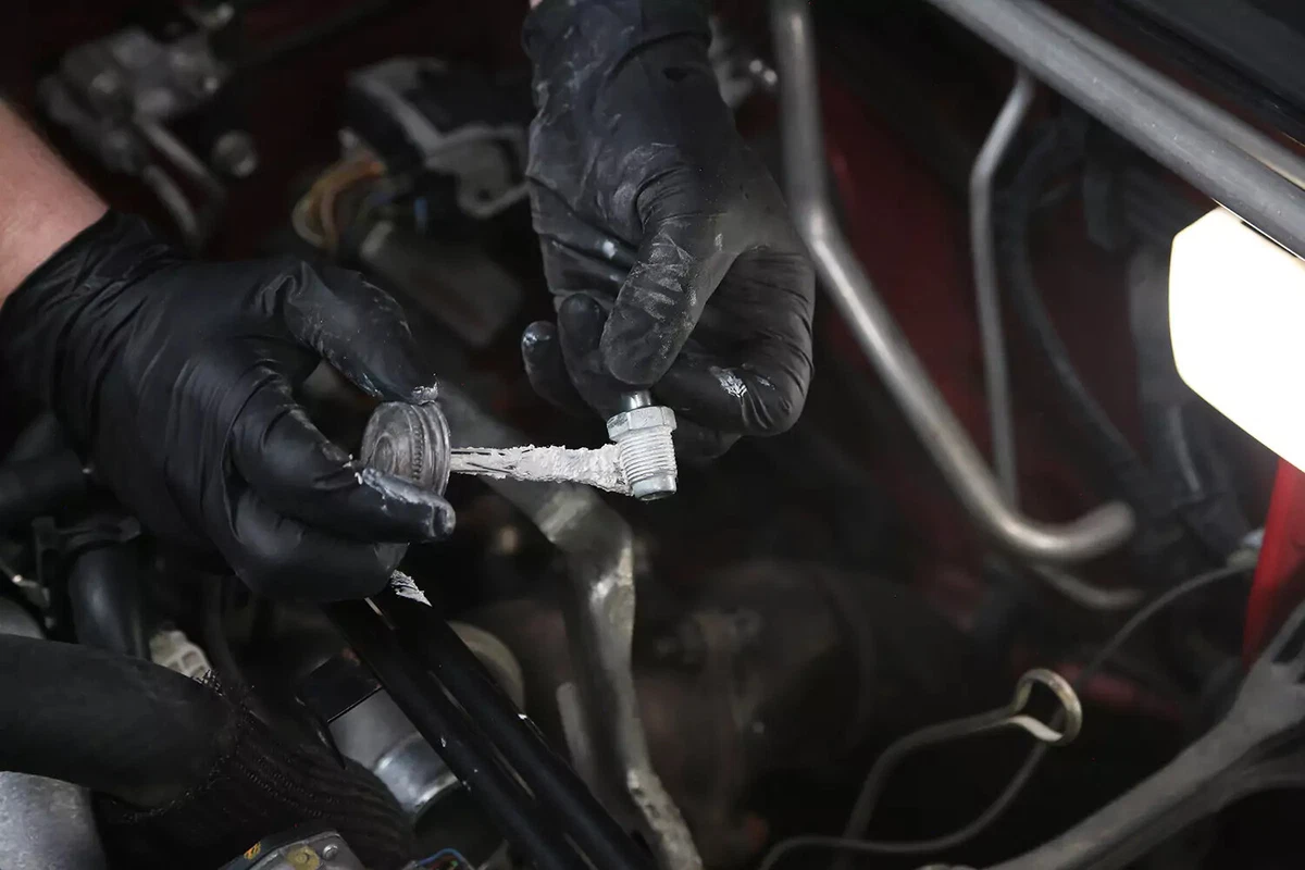 A mechanic wearing black gloves applies sealant to the threads of a PCV valve.