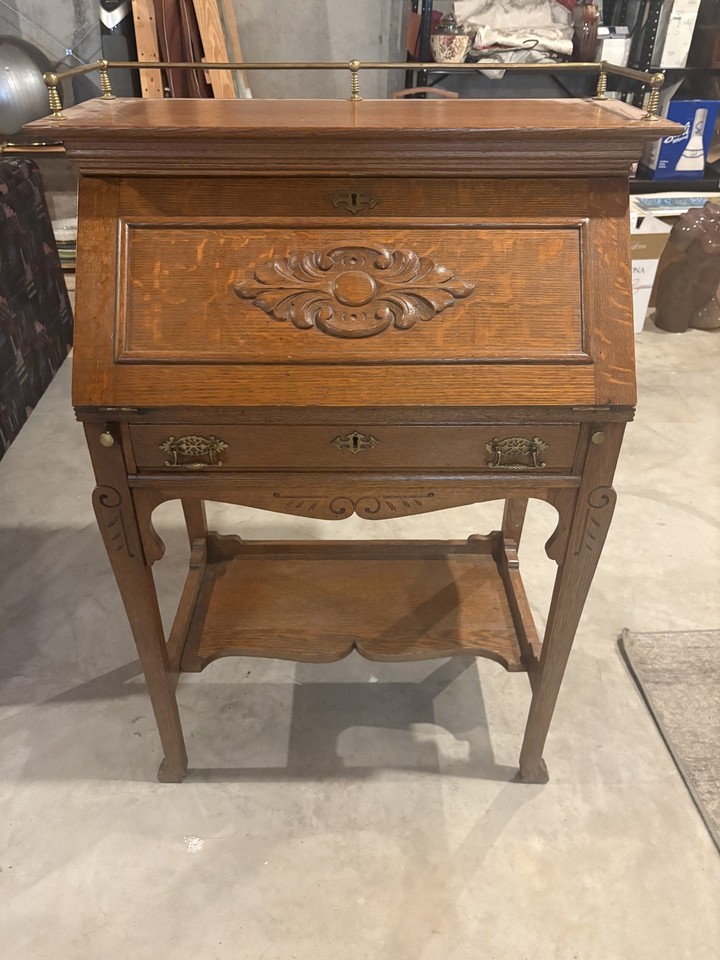 A carved drop-front wooden secretary desk with a brass gallery rail — a genuinely beautiful antique piece that transforms any room into something with actual character.