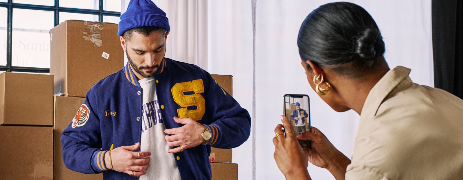 Eine Person posiert und richtet ihre College-Jacke zurecht, während die andere mit einem Smartphone ein Foto macht.