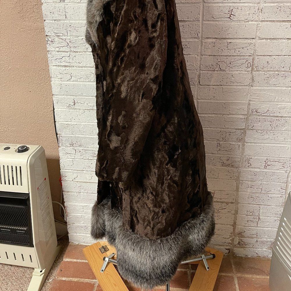 Tall brown fur winter coat with silver-black trim, displayed on a yellow wooden block against a brick wall.