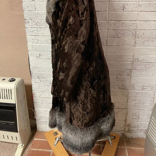 Tall brown fur winter coat with silver-black trim, displayed on a yellow wooden block against a brick wall.