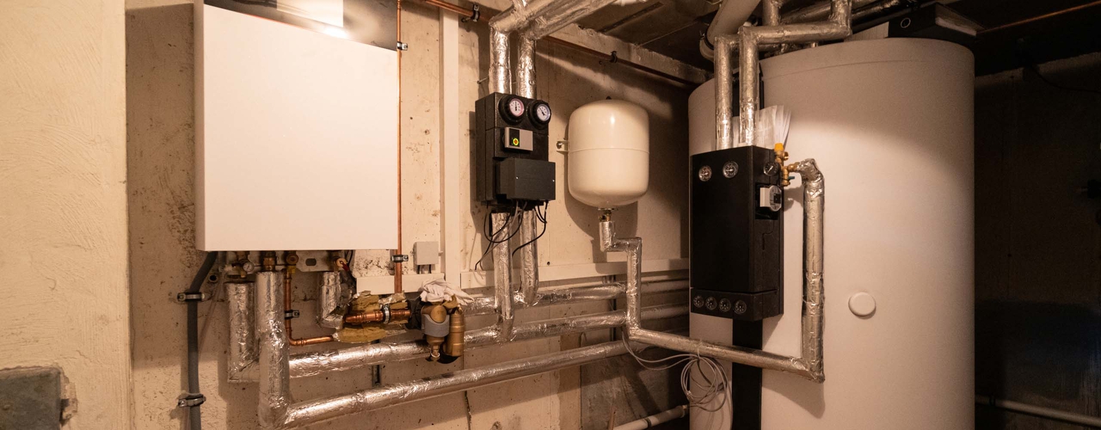 Ein Heizungsraum mit einer modernen Heizungsanlage, bestehend aus einem weißen Wandkessel, einem schwarzen Steuergerät, einem weißen Ausgleichsbehälter und einem großen weißen Warmwasserspeicher