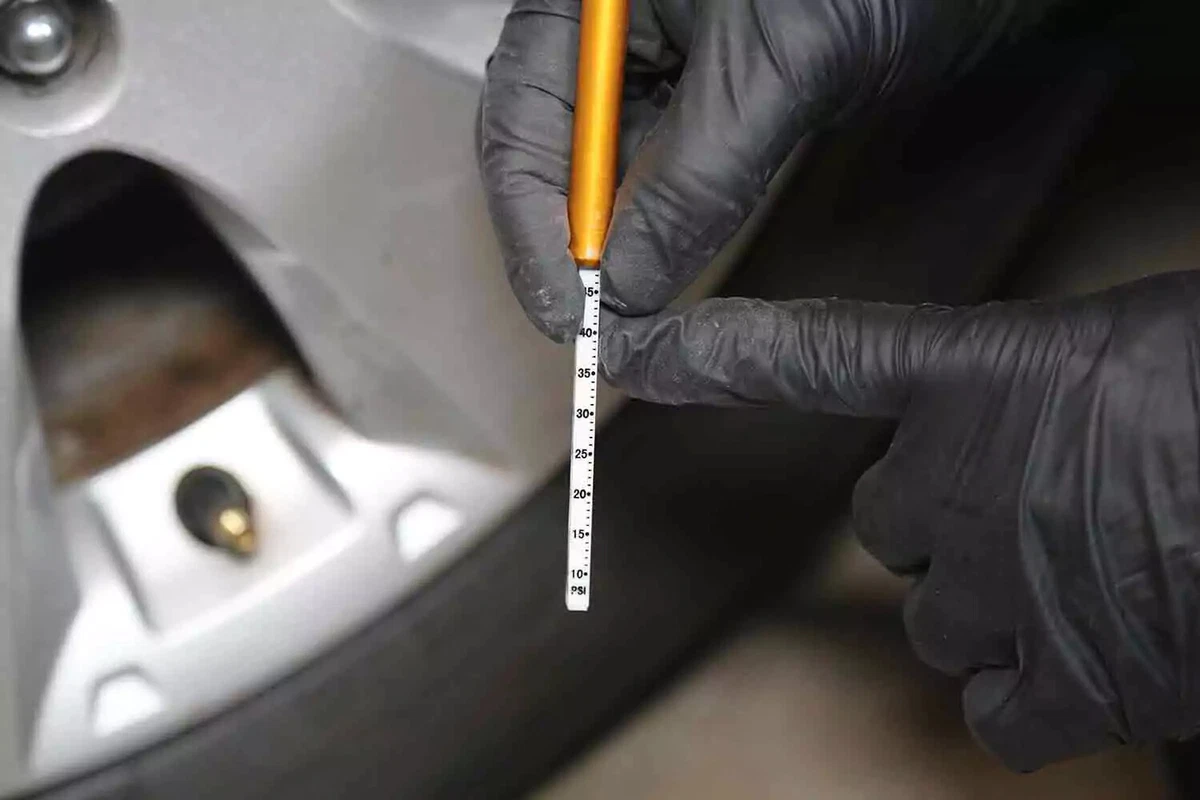 A mechanic wearing black gloves points to the readout on a manual tire pressure gauge.