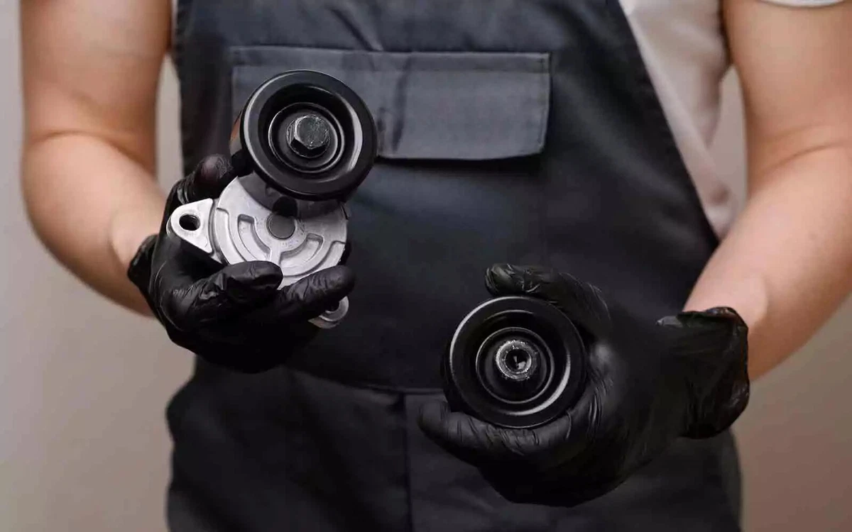A mechanic in overalls, wearing black nitrile gloves, holds two idler pulleys. The pulley on the left is connected to its tensioner.