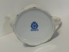 White porcelain teapot with ornate lid, gold rim, and blue floral medallion reading “Winrose Collection Hand Decorated in U.S.A.”