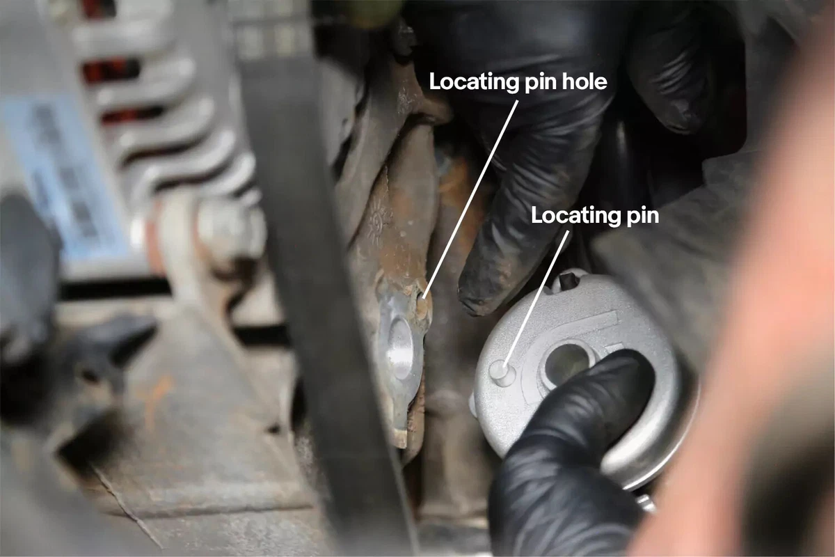 A mechanic wearing black gloves lines up the serpentine belt locating pin with the locating pin hole on the engine block.