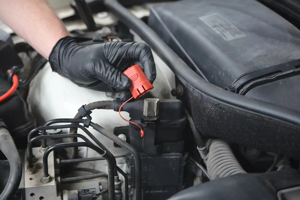 A mechanic wearing a black glove removes the red cap from a car battery's positive post.