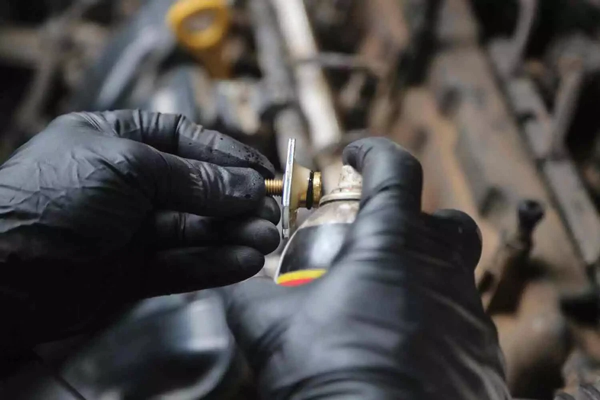 A mechanic wearing black gloves applies lubricant to the o-ring for a fuel injector.