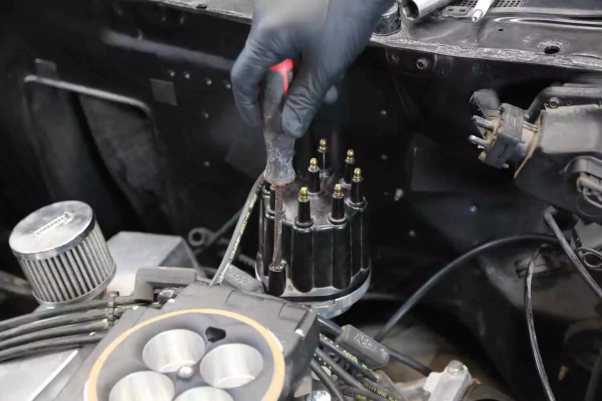 A mechanic wearing a black nitrile glove unfastens a distributor with a screwdriver.
