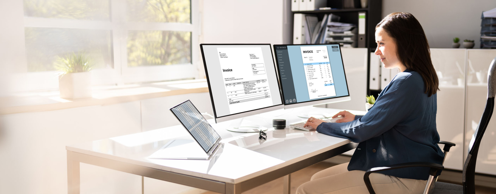 Eine Frau arbeitet in einem modernen Büro an einem Schreibtisch mit zwei großen Monitoren und einem Laptop.