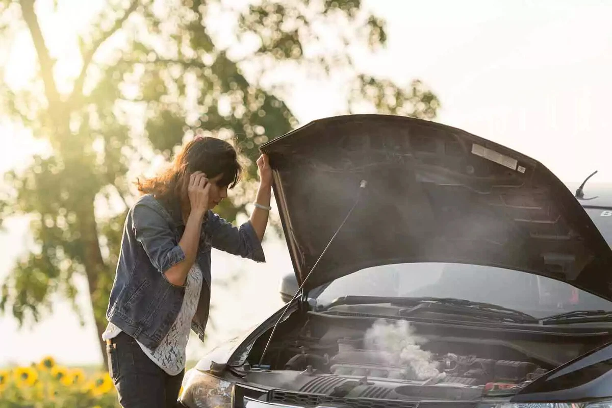 A driver on her cellphone, holding her car’s open hood, as the radiator steams and the sun shines through a tree in the background.