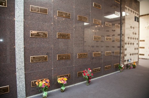 Bronze plaques with names on dark marble wall, vases of flowers at base, in indoor mausoleum crypt.