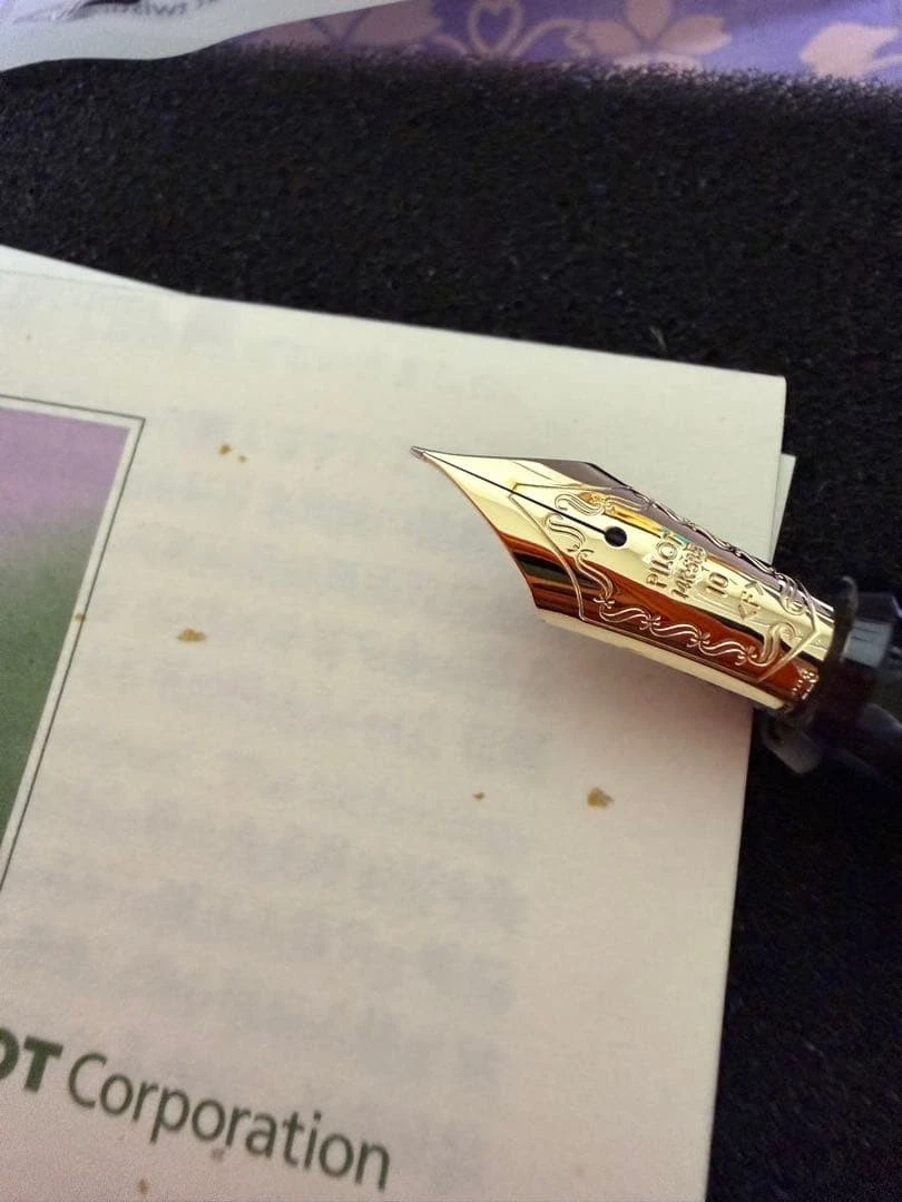 Gold-toned UNUSED Pilot fountain pen with black nib, white body, and ornate scrollwork handle on a marbled surface.