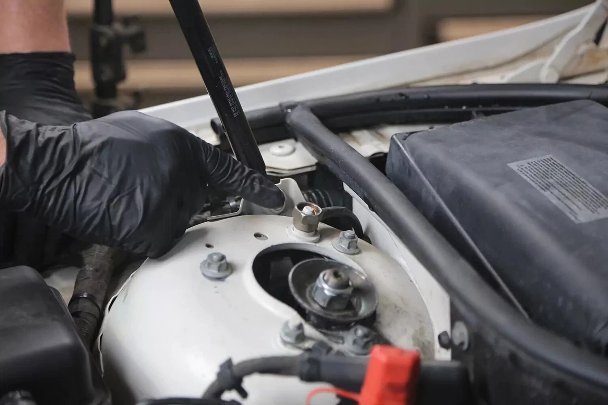 A mechanic wearing a black glove points to a hex nut to be used as a grounding point for jump starting.