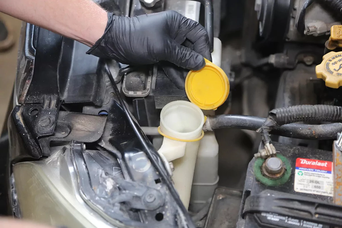 A mechanice wearing a black glove opens the yellow cap on a vehicle's windshield fluid reservoir.