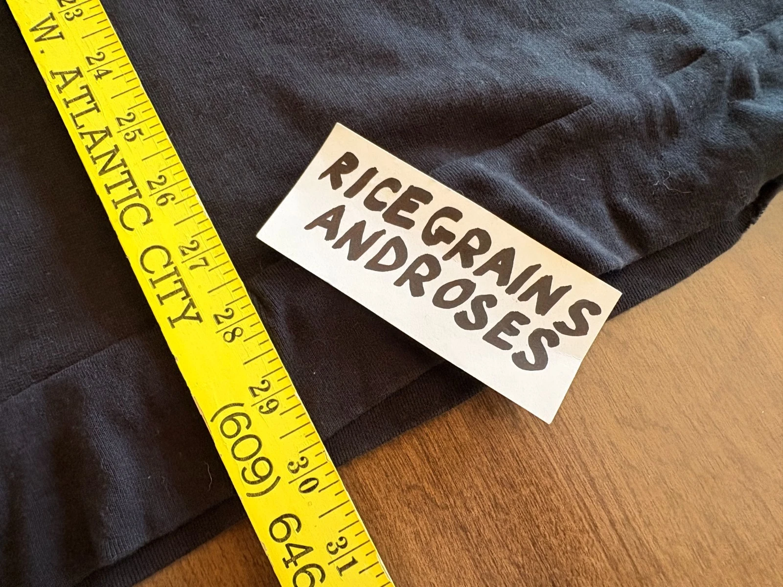 Black crewneck sweater size medium with white label reading “RICE Grains ANDROSES” in yellow text, shown beside a measuring tape.