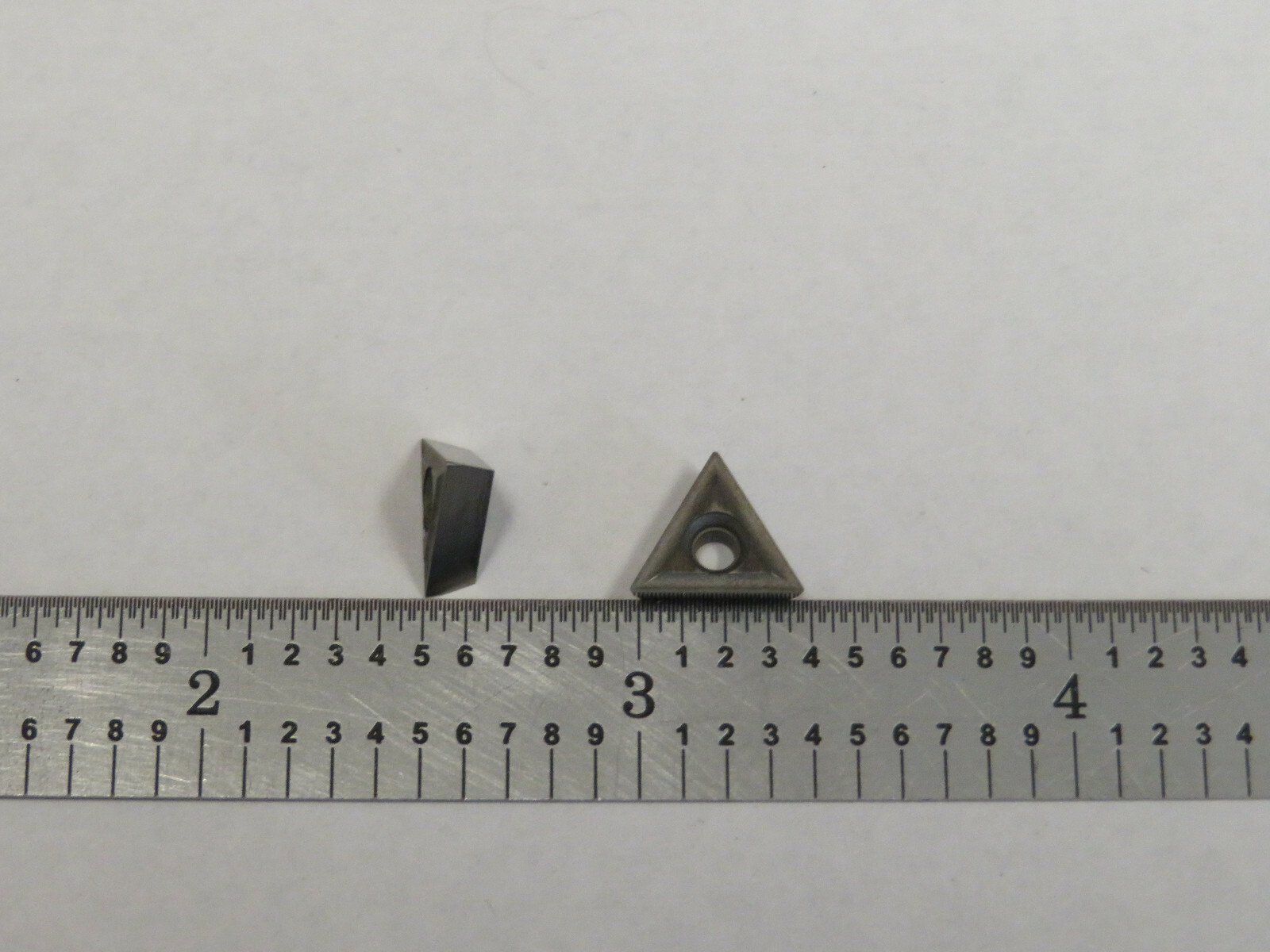 Small metal TT-221 carbide insert with triangular cutting head, shown beside a ruler on a gray surface.