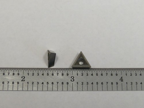 Small metal TT-221 carbide insert with triangular cutting head, shown beside a ruler on a gray surface.