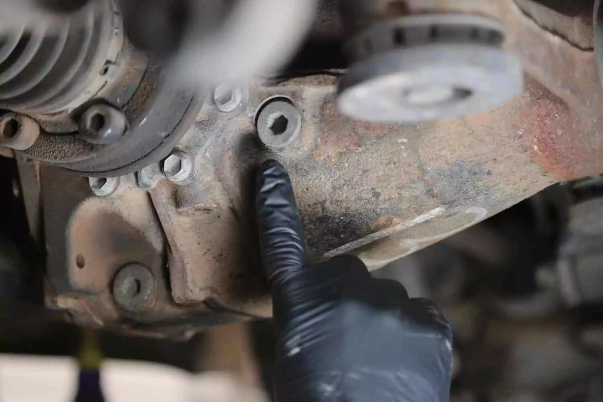A mechanic wearing a black glove points to the fill plug of a vehicle's rear differential.