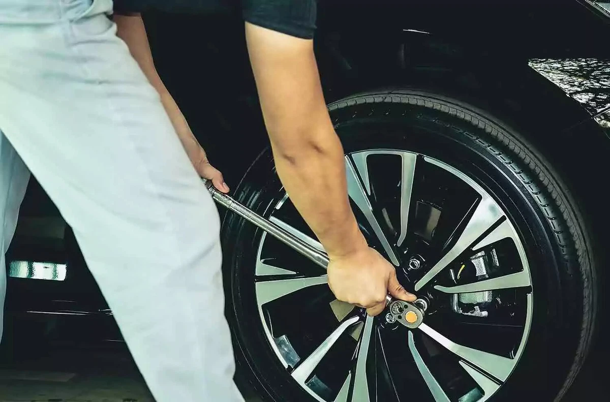 A driver wearing jeans and a black t-shirt tightens a lug nut on an alloy wheel using a torque wrench.