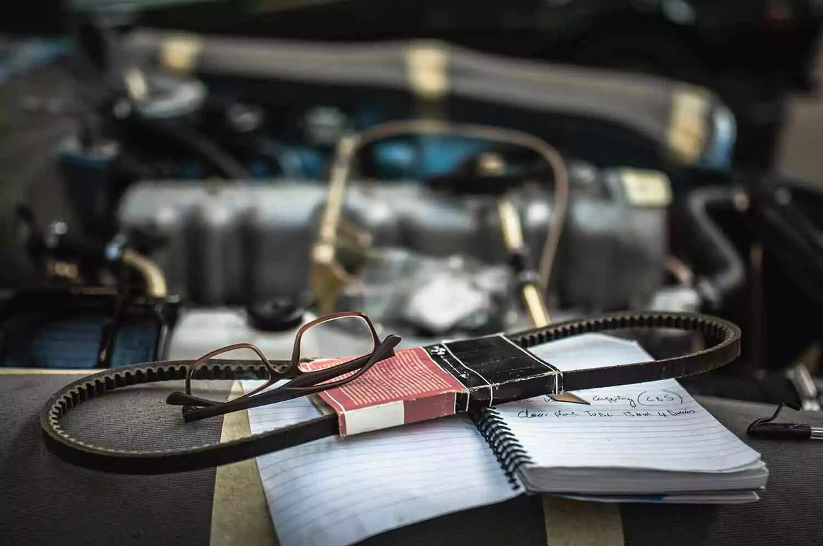 A set of eyeglasses sit on top of a new drive belt and a notebook, on the edge of a vintage car’s engine compartment.