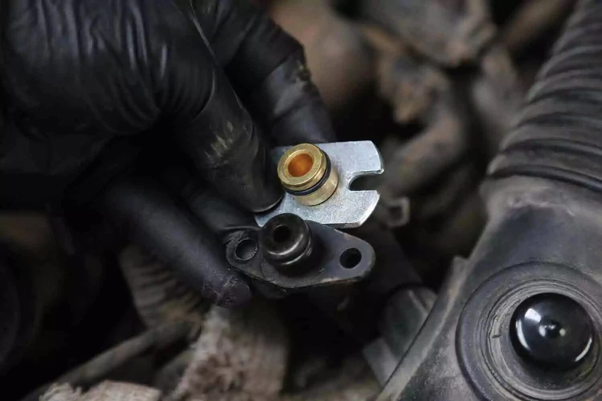A mechanic wearing a black glove compares a new silver fitting to an old black one.