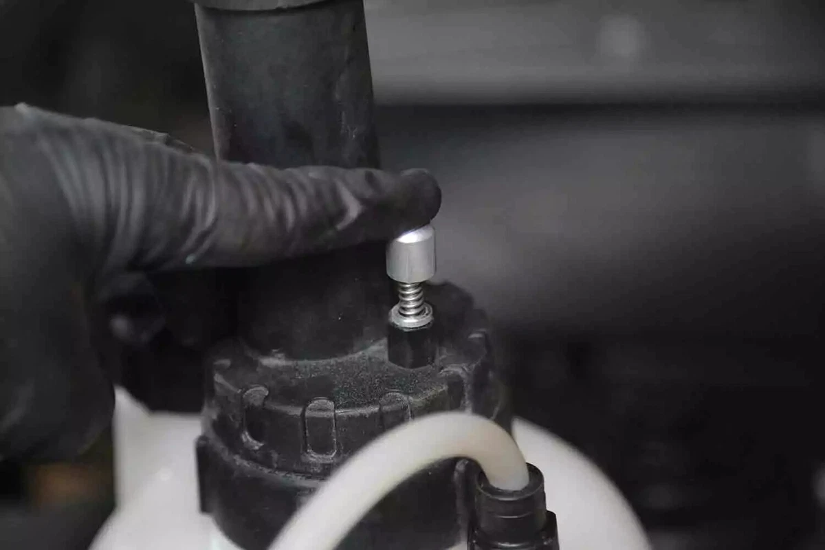 A mechanic wearing a black glove presses the purge valve on a brake bleeder tank.