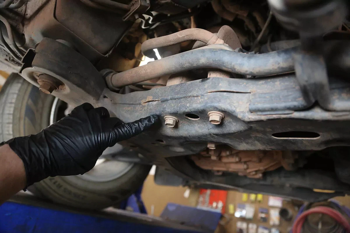 A mechanic with a black glove points at two bolts holding the transmission mount in place.