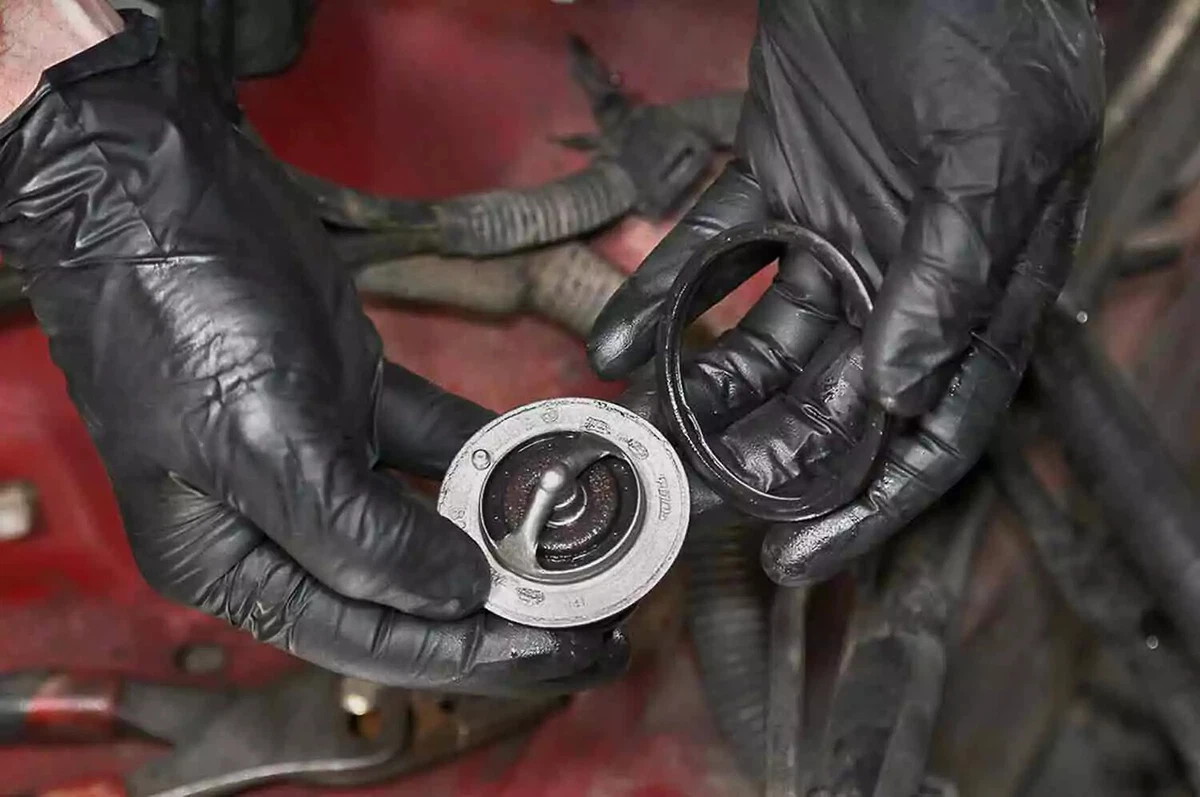 A mechanic wearing black nitrile gloves, holding a used thermostat and gasket in front of a car with a red engine bay.