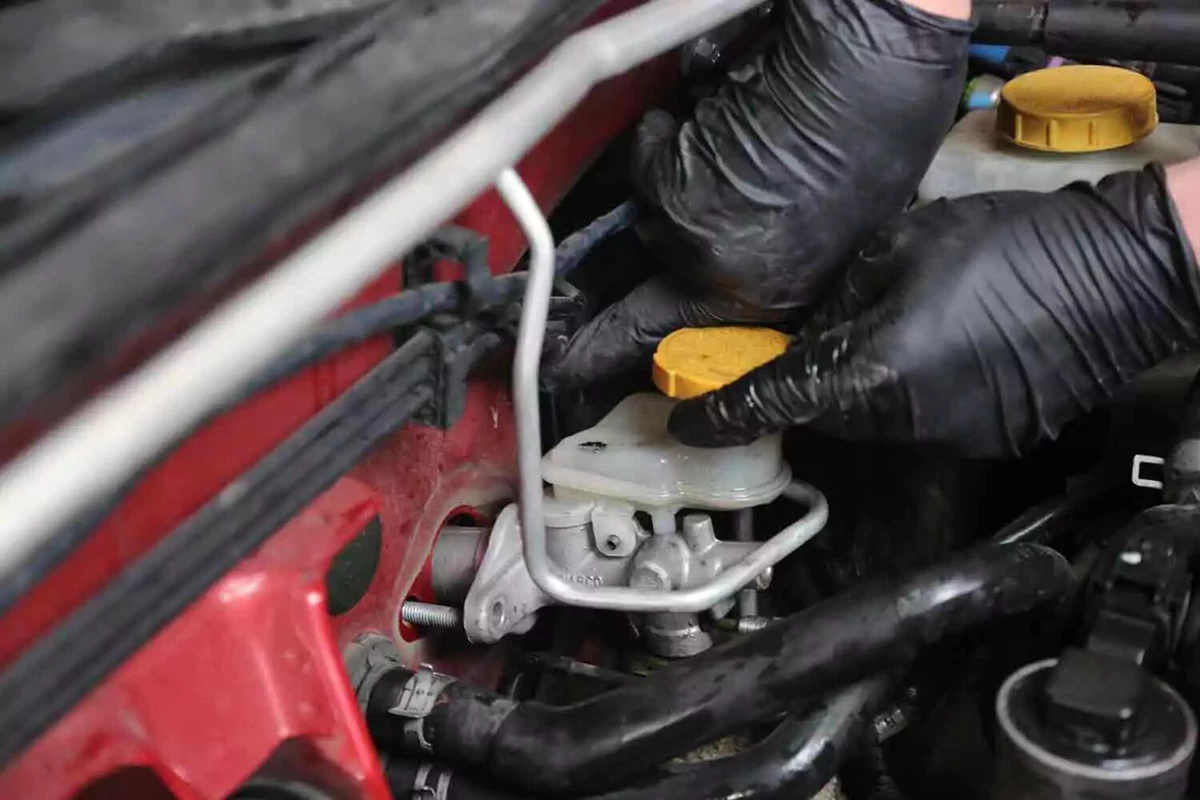 A mechanic wearing black gloves twists the clutch master to remove it from the firewall.