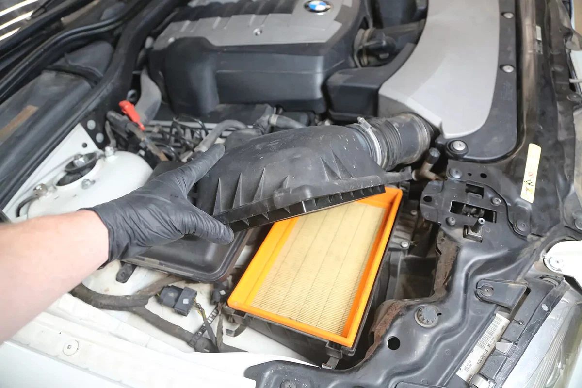 A mechanic wearing a black glove opens the vehicle's engine air filter box and reveals the engine air filter inside.