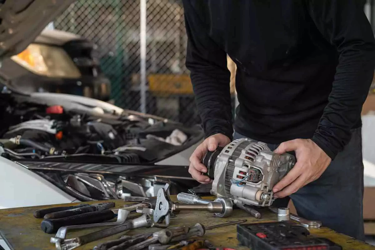 Mechanic holding a new alternator, standing next to a car with an open hood and a work table full of hand tools.