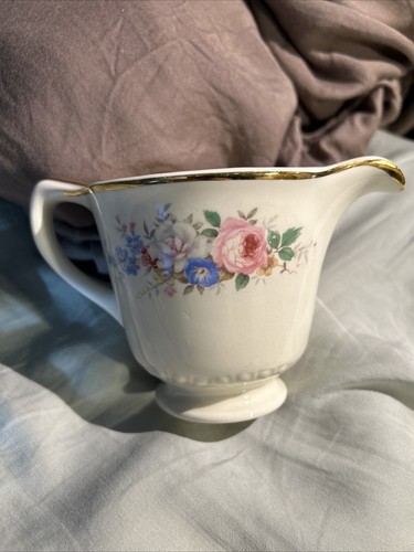 White ceramic pitcher with gold rim, pink rose floral print, and blue hydrangea accents on a bed with gold fabric.