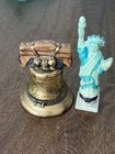 Two miniature Liberty Bell statues, one gold-toned with blue robe, one white with gold accents, on wooden surface.